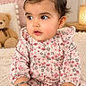 baby, child, infant, portrait, sitting, floral_dress, pink_clothing, hair_bow, wide_eyes, looking_away, indoors, cozy, blanket, teddy_bear, flowers, lamp, soft_lighting, nursery, carpet, happy