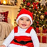 Myra joined the competition — help win amazing prizes! baby, child, santa_costume, christmas, christmas_tree, holiday, smiling, sitting, portrait, indoor, living_room, carpet, wrapped_gifts, ornaments, red_bow, snowflake_ornament, festive, warm_lighting, infant, family_home