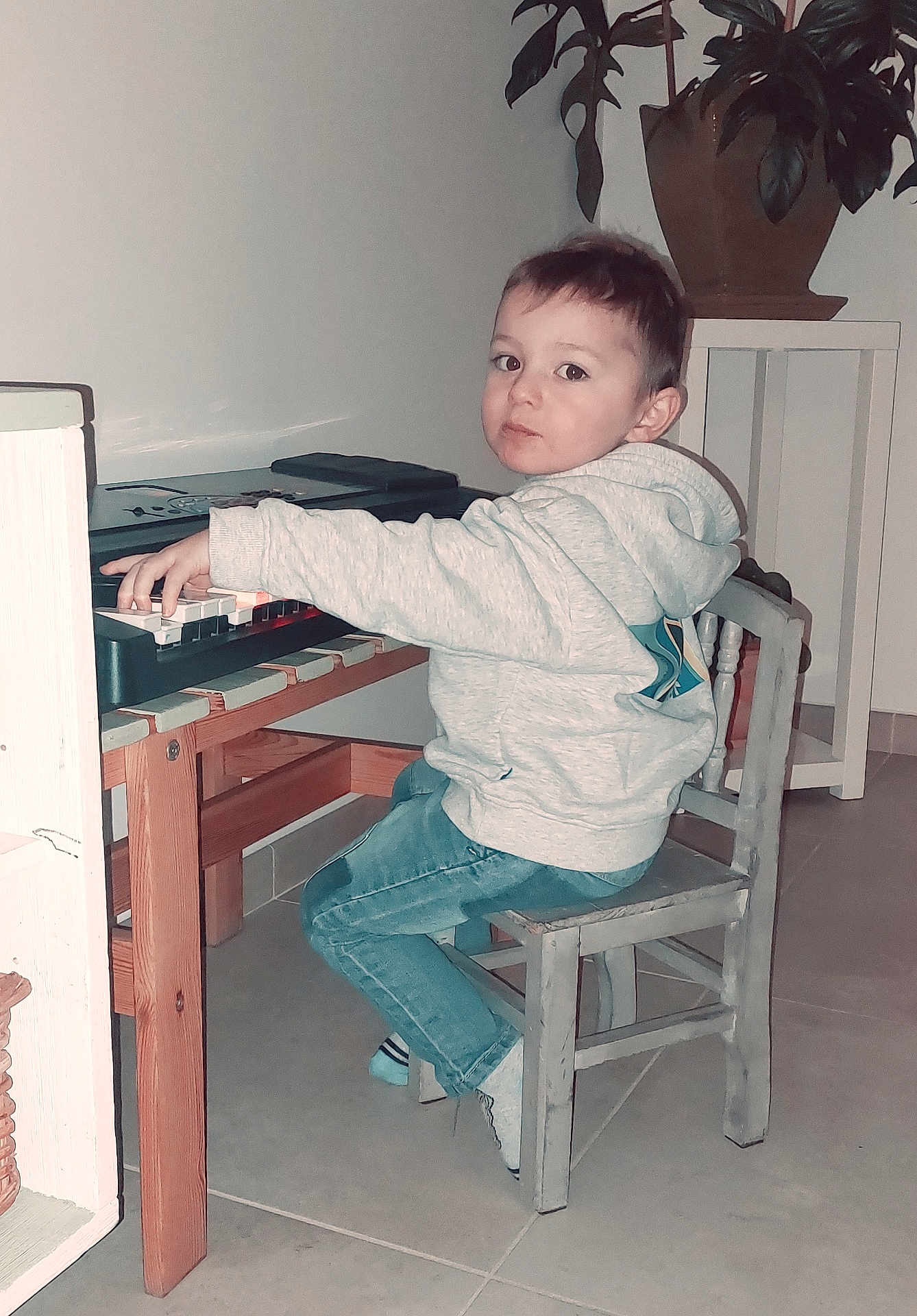 Joris participe au concours pour gagner de l'argent avec cette photo : child, indoor, keyboard, piano, music, playing, hoodie, jeans, chair, wooden_furniture, plant, potted_plant, floor_tiles, curious_expression, young_child, small_chair, casual_clothing, person, musical_instrument, home