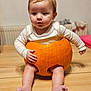 baby, pumpkin, child, cute, infant, orange, table, wooden_surface, indoors, white_clothing, legs, feet, hands, face, curly_hair, sitting, soft_light, holiday, autumn, funny
