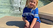 Hadley-Rae joined the competition — help win amazing prizes! blonde_hair, blue_shirt, casual_clothing, child, daytime, fountain, grass, nature, outdoor, park, playful, recreation, shoes, sitting, smiling, stone_wall, sunny, trees, water_splash, young_child