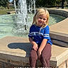 Hadley-Rae joined the competition — help win amazing prizes! blonde_hair, blue_shirt, casual_clothing, child, daytime, fountain, grass, nature, outdoor, park, playful, recreation, shoes, sitting, smiling, stone_wall, sunny, trees, water_splash, young_child