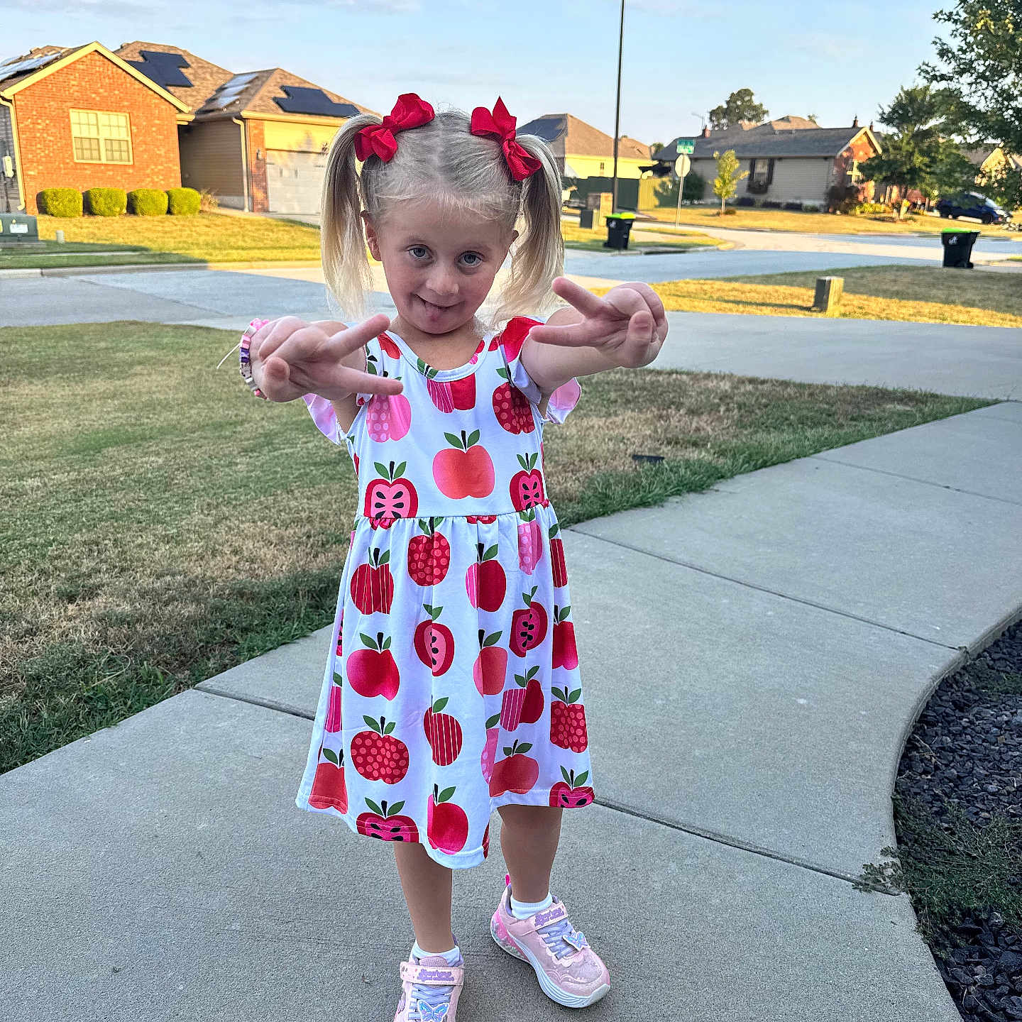 Hadley-Rae joined the competition — help win amazing prizes! child, girl, pigtails, red_bows, dress, apples, sneakers, sidewalk, suburb, neighborhood, grass, street, houses, daylight, sky, playful, peace_sign, smile, outdoor, person