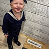 Cody is registered to the contest to win money with this photo: child, toddler, smiling, sailor_outfit, hat, floor, vent, wooden_wall, indoor, portrait, footwear, black_shoes, happy, standing, clothing, cute, person, young_child, fashion, casual