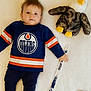 baby, blue_clothing, child, cute, eagle, hockey_stick, indoor, lying_down, orange_stripes, plush_toy, portrait, small_hockey_puck, smiling, snowflake, sock, soft_texture, sports_jersey, toy, white_blanket, winter_theme