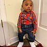 toddler, child, standing, red_shirt, plaid, jeans, white_sneakers, indoor, door, wall, curious, young_child, fashion, casual_wear, floor, rug, small_person, portrait, cute, person