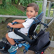 Pablo a rejoint le concours — aidez-le/la à gagner de superbes lots ! child, tricycle, outdoor, smiling, greenery, trees, metal_railing, shorts, white_shirt, sneakers, footrest, seat, handlebars, park, pathway, happy, person, daylight, nature, play