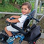 child, tricycle, outdoor, smiling, greenery, trees, metal_railing, shorts, white_shirt, sneakers, footrest, seat, handlebars, park, pathway, happy, person, daylight, nature, play