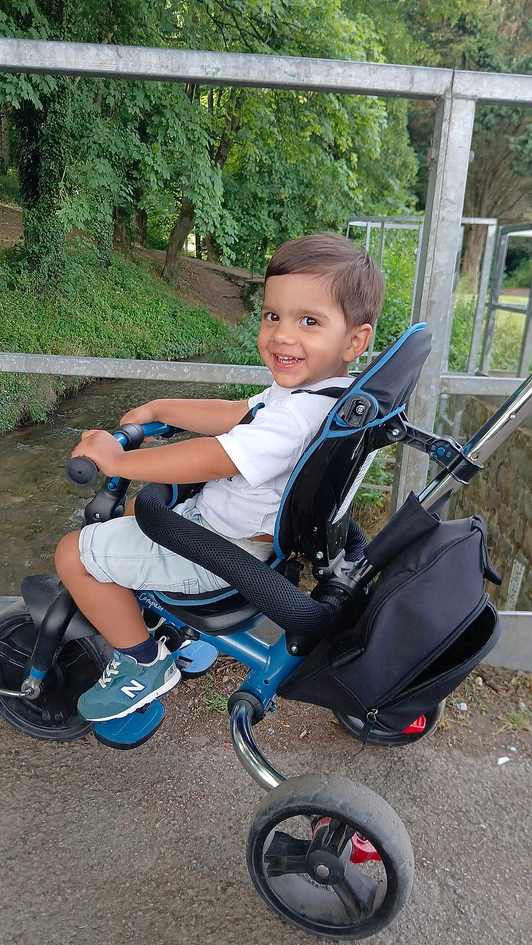 Pablo a rejoint le concours — aidez-le/la à gagner de superbes lots ! child, tricycle, outdoor, smiling, greenery, trees, metal_railing, shorts, white_shirt, sneakers, footrest, seat, handlebars, park, pathway, happy, person, daylight, nature, play