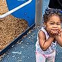 blue, child, clothing, cute, daylight, face, hair_bows, hand_on_face, legs, outdoor, patterned_pants, person, playground, playground_equipment, purple, smiling, standing, toddler, white, wood_chips