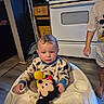 baby, child, infant, high_chair, mickey_mouse, stuffed_toy, toy, kitchen, stove, appliances, indoor, sitting, portrait, curious, clothing, pajamas, smiling, wood_floor, home, family