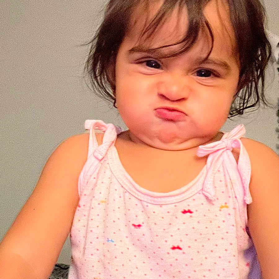 Maryam joined the competition — help win amazing prizes! child, toddler, baby, portrait, indoors, pink_outfit, dress, sitting, facial_expression, pouting, puffed_cheeks, brown_hair, shoulder_length_hair, looking_at_camera, closeup, home, wall, ceiling, carpet, humorous