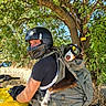 person, dog, motorcycle, helmet, goggles, backpack, gloves, tree, outdoor, sunlight, yellow, leaves, nature, animal, pet, riding, transportation, adventure, protective_gear, summer