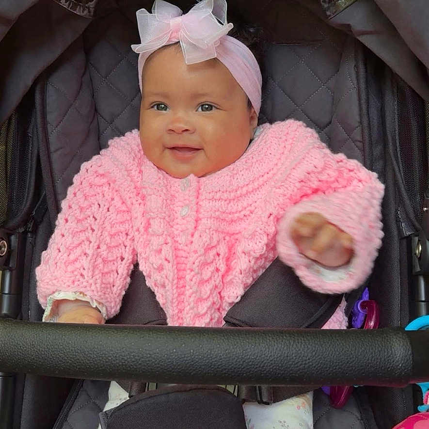 Theia is registered to the contest to win money with this photo: baby, bow, child, closeup, cozy, cute, face, happy, headband, indoor, infant, patterned_pants, pink_sweater, portrait, seated, smiling, socks, stroller, toy, warm_clothing