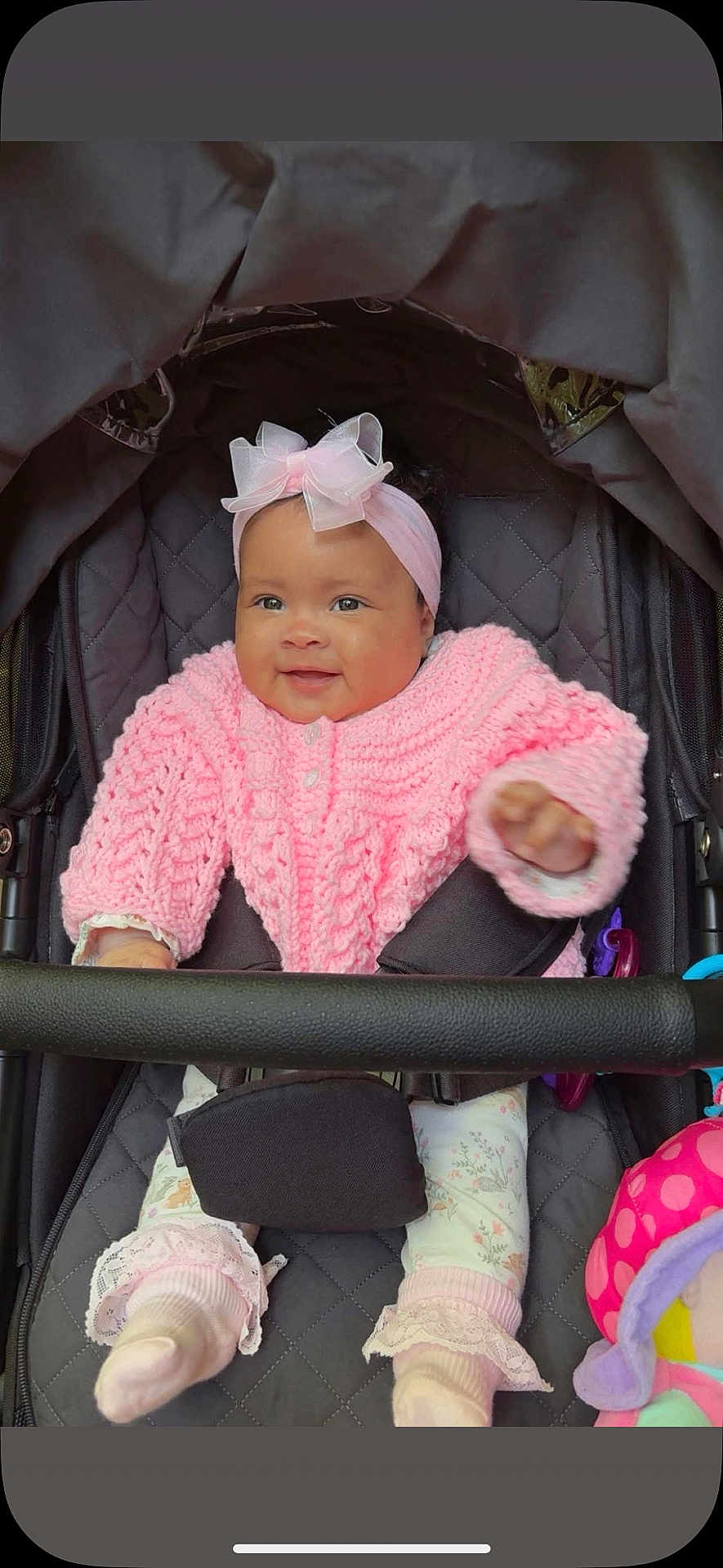 Theia is registered to the contest to win money with this photo: baby, child, smiling, pink_sweater, headband, bow, stroller, toy, cute, infant, cozy, warm_clothing, socks, patterned_pants, happy, face, portrait, seated, indoor, closeup