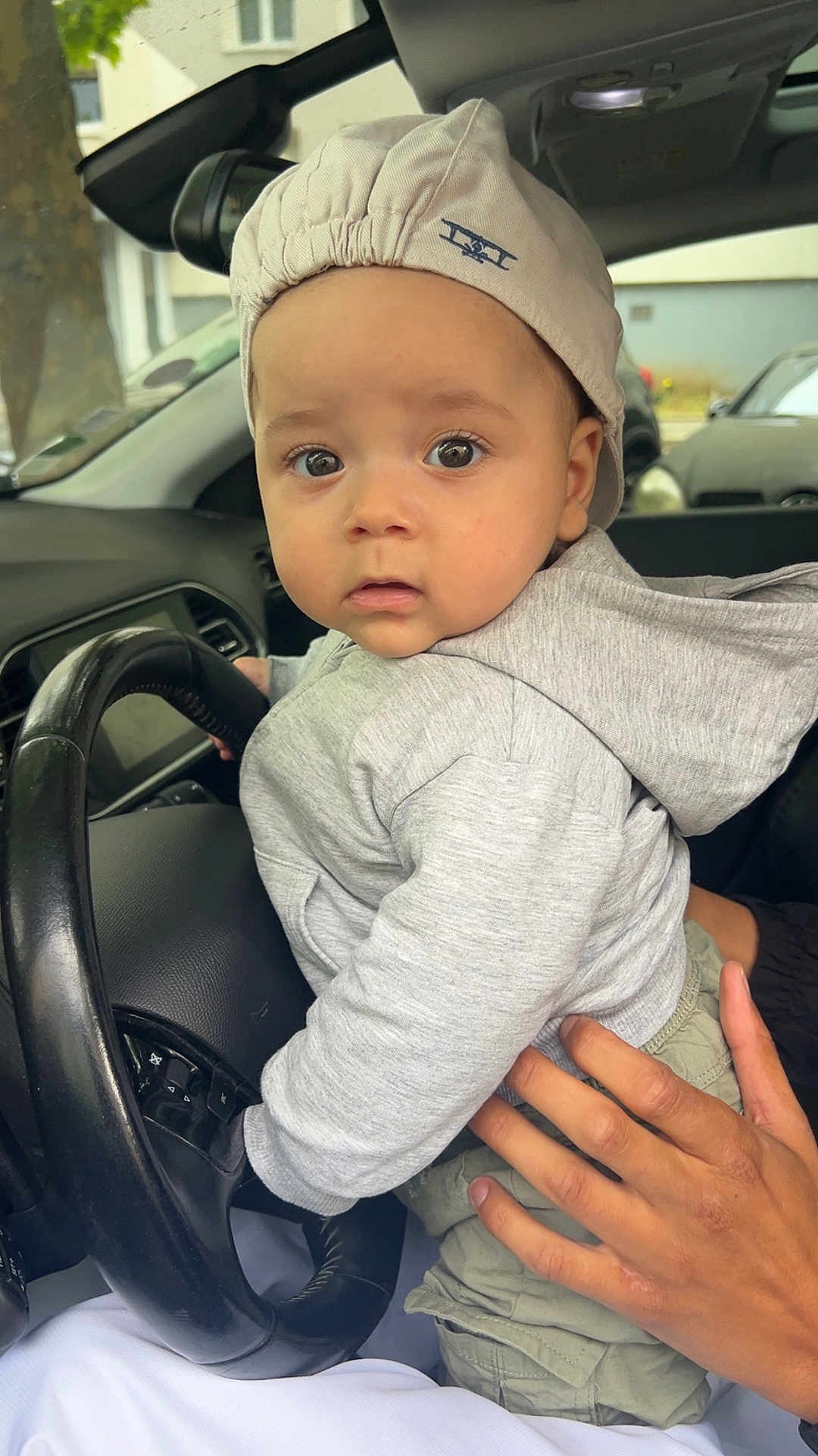 Noah a rejoint le concours — aidez-le/la à gagner de superbes lots ! toddler, child, baby, cap, hoodie, hand, steering_wheel, car_interior, curious, portrait, clothing, seat, driving, vehicle, window, urban, casual, person, cute, indoor