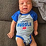 baby, infant, smiling, onesie, blue_clothing, gray_clothing, text_on_clothing, couch, blanket, happy, face, head, skin, arm, hand, leg, foot, indoors, person, baby_clothes