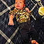 baby, infant, plaid_bedspread, camouflage_shirt, dark_pants, lying_down, barefoot, baby_lotion, clothing, bed, fabric, person, newborn, small_hand, small_feet, casual_wear, indoors, baby_grooming, resting, portrait