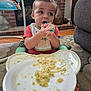 baby, bib, carpet, child, couch, curious, eating, face, floor, food, hands, high_chair, home, indoor, living_room, messy, plate, red_shirt, toy, young