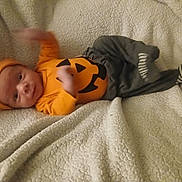 Micha is registered to the contest to win money with this photo: baby, infant, orange_clothing, pumpkin_outfit, hat, blanket, cozy, smiling, hand_motion, striped_sock, gray_pants, cute, lying_down, indoor, soft_texture, young_child, face, headwear, warm_clothing, portrait