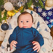 Colt is registered to the contest to win money with this photo: baby, infant, blue_clothing, blanket, christmas_tree, christmas_ornaments, holiday, decorations, face, eyes, hands, indoor, festive, cute, child, smiling, seasonal, cozy, portrait, celebration