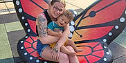 Carmello is registered to the contest to win money with this photo: affection, bench, butterfly_bench, casual_clothing, child, flip_flops, glasses, grass, happy, nature, outdoor, park, playground_surface, shorts, smile, sunlight, tattoo, toddler, trees, woman