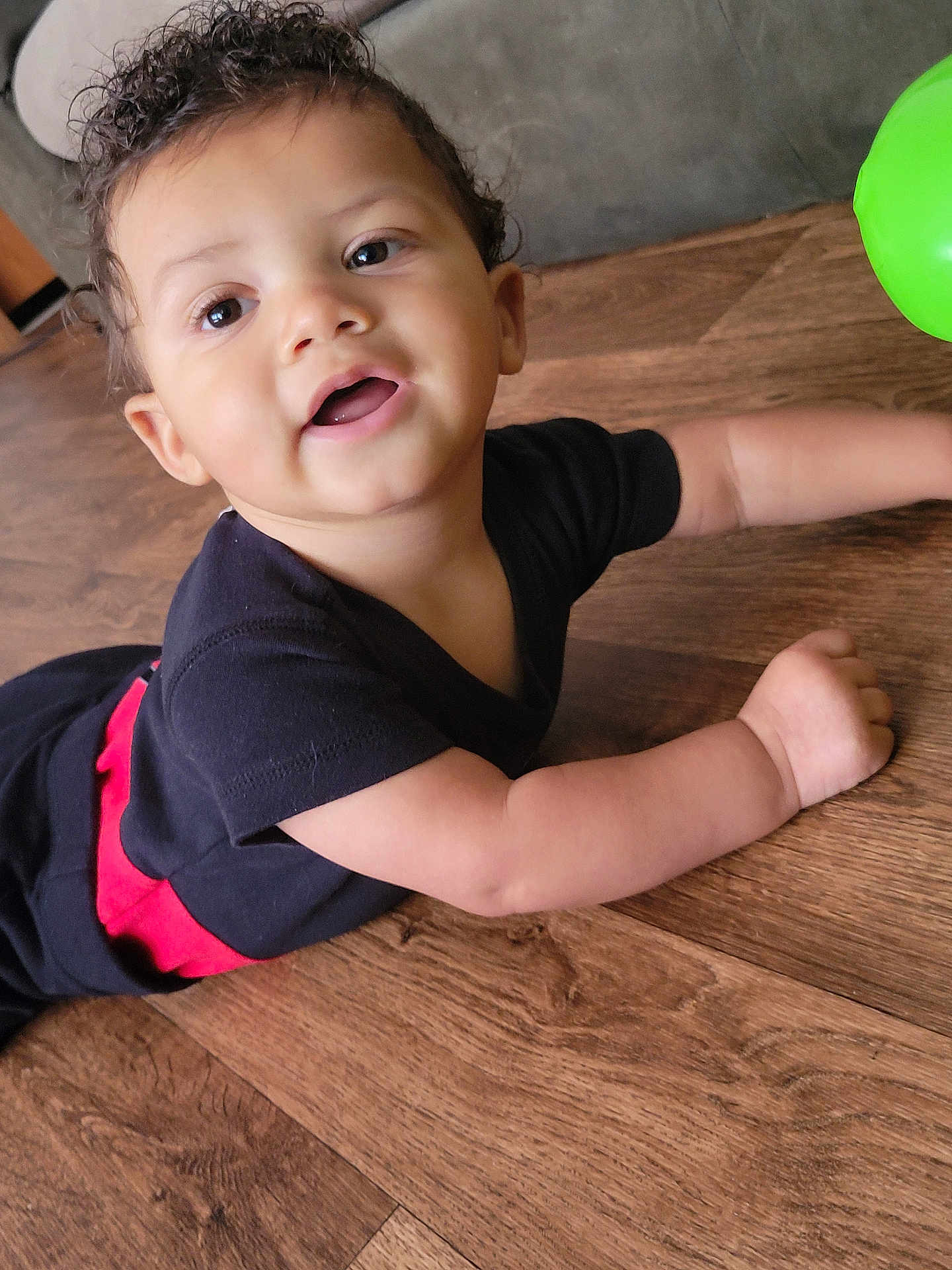 Carmello is registered to the contest to win money with this photo: toddler, child, baby, curly_hair, black_clothing, red_detail, green_ball, wooden_floor, indoor, flooring, smiling, happy, cute, person, skin, face, arm, open_mouth, playful, crawling