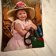 Lillie is registered to the contest to win money with this photo: child, girl, hat, straw_hat, pink_dress, flower, rose, steering_wheel, wooden_fence, american_flag, portrait, vintage, indoor, smile, face, person, marble_surface, photograph, pose, cute