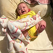 Amy is registered to the contest to win money with this photo: baby, smiling, blanket, yellow_clothing, cushion, indoor, happy, infant, lying_down, cute, child, soft_fabric, cozy, head, feet, person, portrait, comfort, wrapped, playful