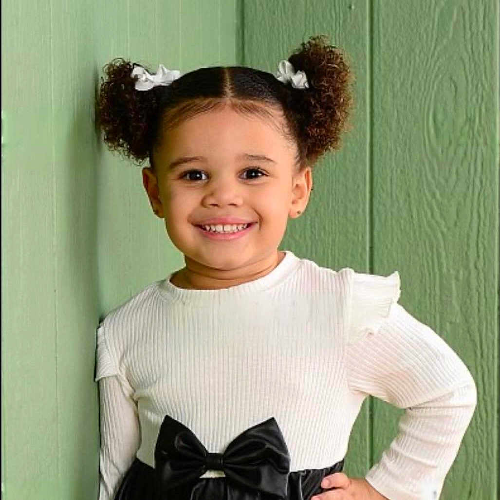 Giovanna Gilbert is registered to the contest to win money with this photo: black_skirt, bow, child, confident, cute, fashion, girl, green_wall, hand_on_hip, happy, indoor, person, pigtails, portrait, smiling, standing, textured_wall, white_bows, white_top, young