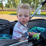 Carter is registered to the contest to win money with this photo: basketball_court, blanket, blue, casual_clothing, child, curly_hair, daytime, expression, face, gray, green, outdoor, park, playground, sippy_cup, stroller, summer, sunlight, toddler, toy
