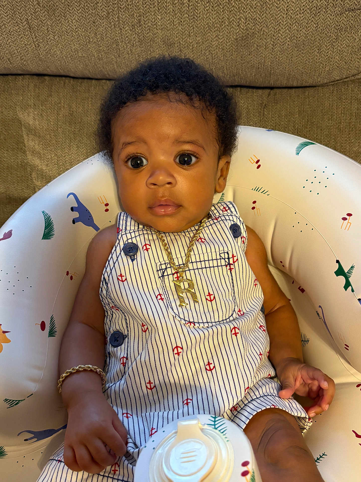 Rasheed is registered to the contest to win money with this photo: baby, infant, child, romper, gold_bracelet, gold_necklace, baby_seat, animal_prints, curly_hair, wide_eyes, striped_clothing, indoor, cute, sitting, portrait, accessories, clothing, person, closeup, baby_toy