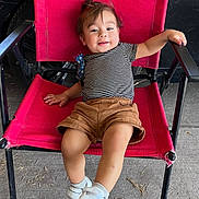 Basilio is registered to the contest to win money with this photo: toddler, child, red_chair, striped_shirt, brown_shorts, white_sneakers, velcro, hair, smile, outdoor, concrete_floor, black_metal_chair_frame, casual_clothing, relaxed_pose, happy, person, legs, footwear, seat, daylight