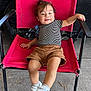 toddler, child, red_chair, striped_shirt, brown_shorts, white_sneakers, velcro, hair, smile, outdoor, concrete_floor, black_metal_chair_frame, casual_clothing, relaxed_pose, happy, person, legs, footwear, seat, daylight