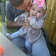 Skylar is registered to the contest to win money with this photo: baby, child, man, outdoor, garden, kiss, pink_bow, floral_dress, happy, smiling, sitting, fingers, headband, person, wooden_fence, chair, table, clothing, family, affection
