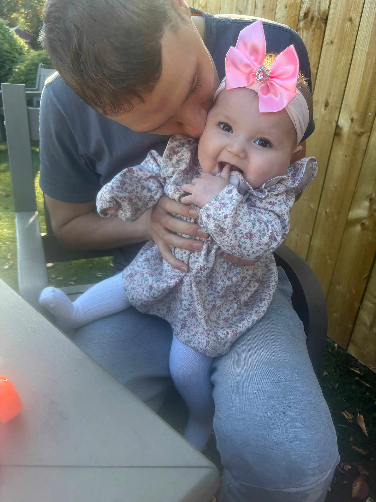 Skylar is registered to the contest to win money with this photo: baby, child, man, outdoor, garden, kiss, pink_bow, floral_dress, happy, smiling, sitting, fingers, headband, person, wooden_fence, chair, table, clothing, family, affection