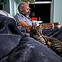 man, cat, tabby_cat, blanket, mug, lamp, window, couch, beard, gray_hair, indoor, relaxing, seated, side_view, furniture, home, casual_clothing, evening, domestic, pet