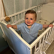 Elio participe au concours pour gagner de l'argent avec cette photo : toddler, child, crib, blue_shirt, smile, indoor, nursery, curtains, wooden_floor, baby, gripping, standing, face, person, furniture, home, knitwear, cute, young_child, happy