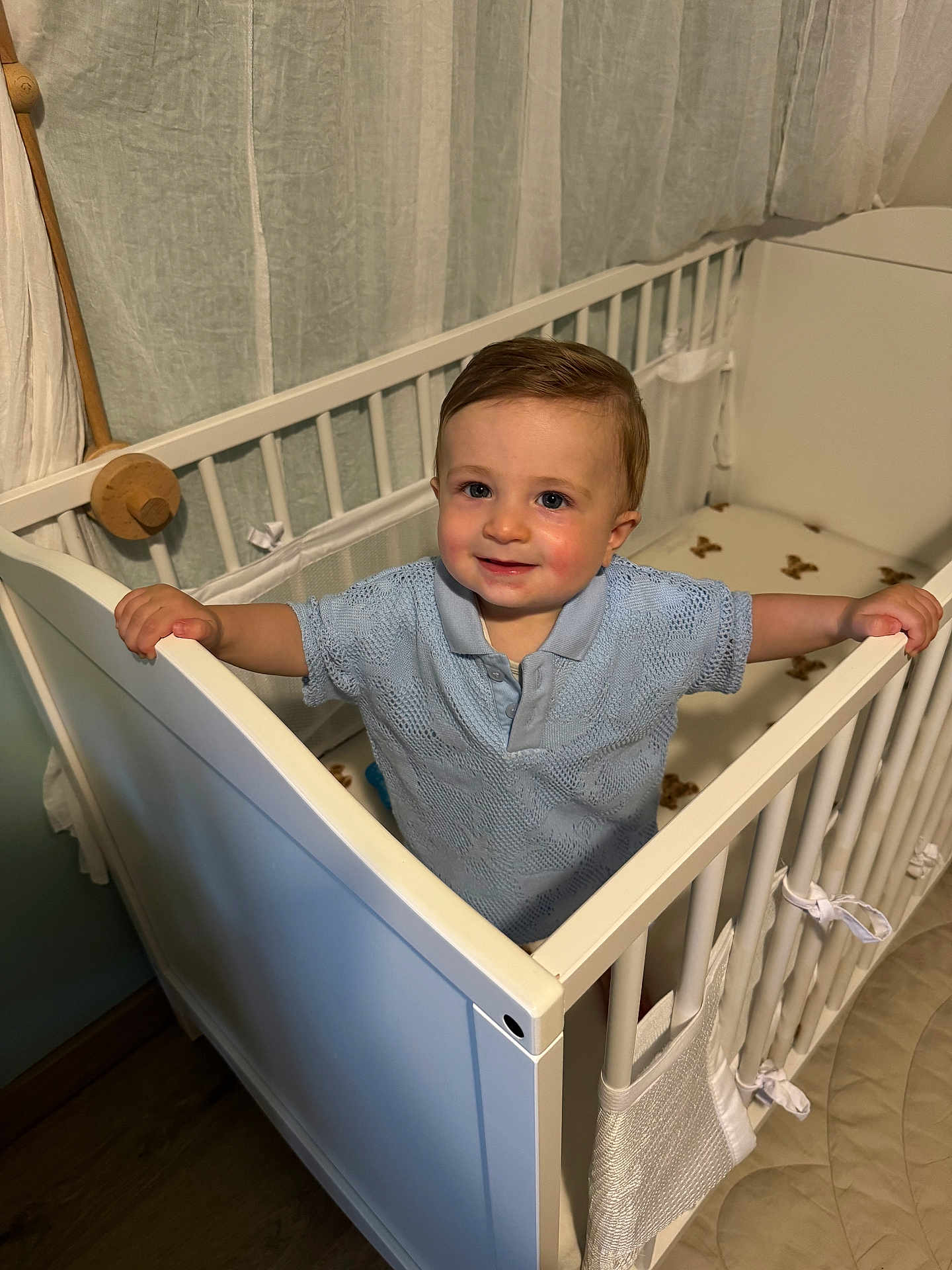 Elio participe au concours pour gagner de l'argent avec cette photo : toddler, child, crib, blue_shirt, smile, indoor, nursery, curtains, wooden_floor, baby, gripping, standing, face, person, furniture, home, knitwear, cute, young_child, happy