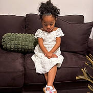 Yetzali is registered to the contest to win money with this photo: calm, carpet, child, couch, crossed_legs, curly_hair, cute, decor, furniture, girl, home, indoor, living_room, looking_down, pillow, portrait, shoes, sitting, table, white_dress