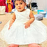 baby, child, dress, white_dress, headband, bow, barefoot, sitting, sparkly_surface, indoor, people_in_background, curious, portrait, cute, child_fashion, celebration, floor, person, face, event