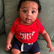Zaylen is registered to the contest to win money with this photo: baby, child, curly_hair, red_shirt, fourth_of_july, navy_shorts, couch, green_couch, texture, sitting, portrait, indoors, cute, wide_eyes, expression, infant, person, clothing, casual, young