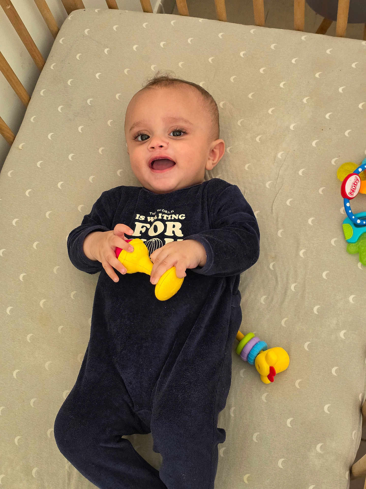 Maïwell participe au concours pour gagner de l'argent avec cette photo : baby, infant, crib, mattress, moon_pattern, onesie, toy, rattle, yellow_toy, smiling, happy, lying_down, playtime, nursery, bedding, face, eyes, hands, wooden_rail, cute