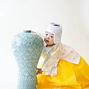 Tay is registered to the contest to win money with this photo: toddler, child, hanbok, traditional_clothing, yellow_skirt, white_hat, ceramic_vase, blue_vase, ornate, patterned, portrait, cute, playful, studio_shot, white_background, asian_child, cultural, smiling, young_child, celebration