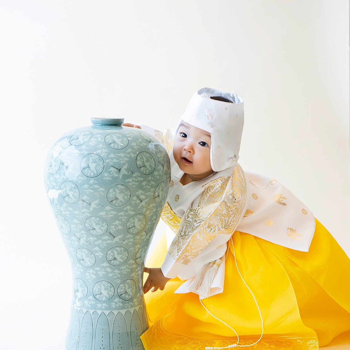 Tay is registered to the contest to win money with this photo: toddler, child, hanbok, traditional_clothing, yellow_skirt, white_hat, ceramic_vase, blue_vase, ornate, patterned, portrait, cute, playful, studio_shot, white_background, asian_child, cultural, smiling, young_child, celebration