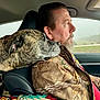 man, dog, car, seat, jacket, camouflage, brindle, headrest, patterned_bag, window, side_profile, beard, driving, pet, resting, shoulder, interior, seatbelt, travel, road