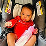 Emonnie is registered to the contest to win money with this photo: baby, infant, child, car_seat, sitting, red_outfit, bottle, baby_bottle, white_socks, curly_hair, wide_eyes, face, hands, legs, indoor, vehicle, safety_labels, portrait, reaching, photograph