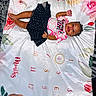 baby, infant, child, lying_down, smiling, portrait, floral_blanket, milestone_blanket, bed, indoor, clothing, skirt, tshirt, barefoot, dark_hair, happy, photography, partial_person, patterned_fabric, family