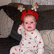 Lottie is registered to the contest to win money with this photo: baby, infant, onesie, strawberry_pattern, red_headband, gingerbread_men, couch, pillow, indoor, cute, child, face, blue_eyes, rosy_cheeks, holiday_accessory, seated, plush, comfort, curious, portrait