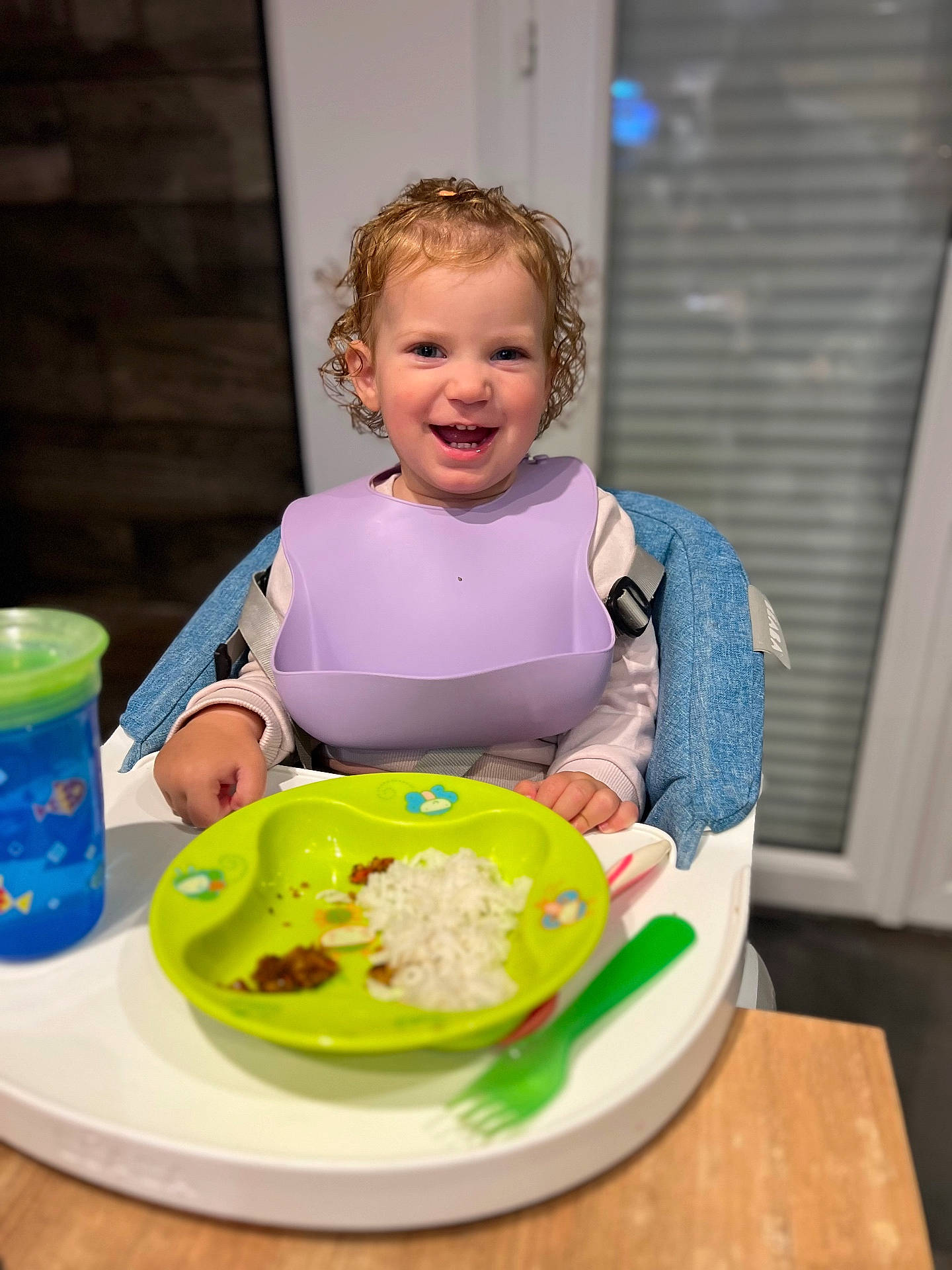 baby, bib, child, curly_hair, eating, food, fork, happy, high_chair, indoor, meal_time, person, plate, rice, sippy_cup, smile, spoon, table, toddler, utensils